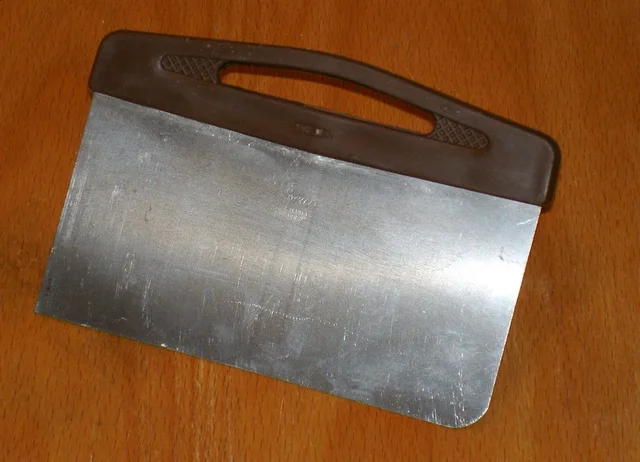 Rectangular metal bench scraper with a dark handle on a wooden surface