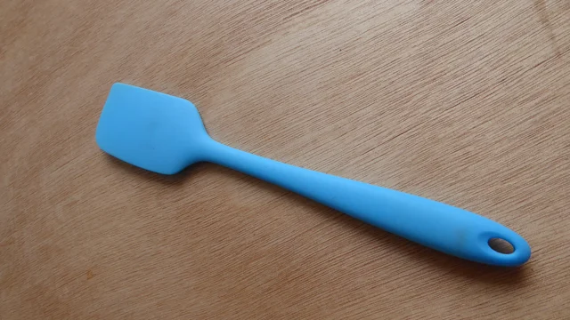 Blue scraping spatula with a broad asymmetrical head and hanging hole on a wooden surface
