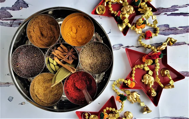 Open round masala box with seven spice-filled metal cups and a spoon