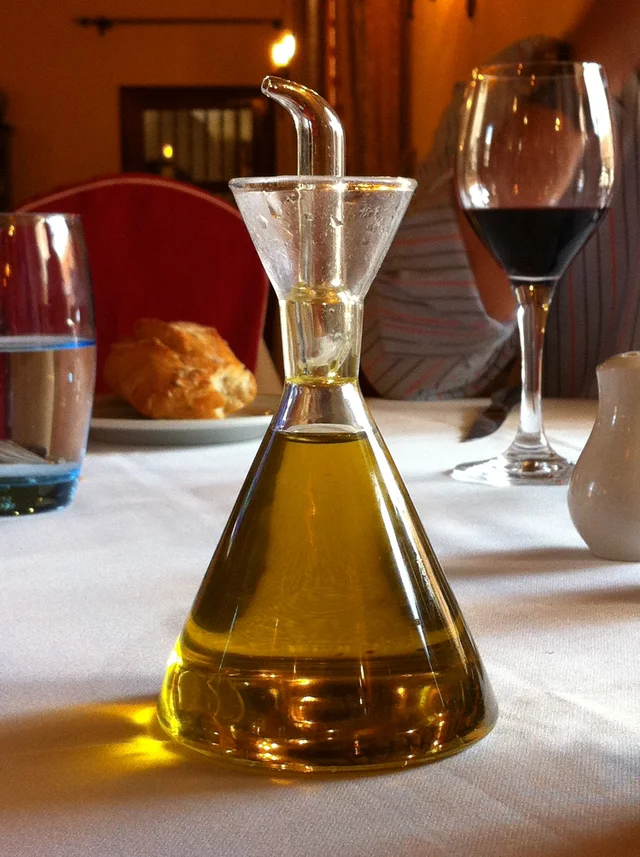 Clear glass oil dispenser with a conical base and curved pouring spout on a dining table