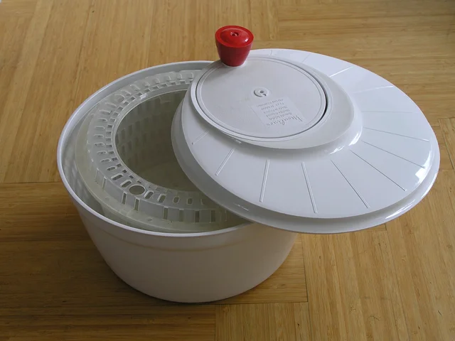White salad spinner with its lid set aside to reveal the inner basket and a red crank knob