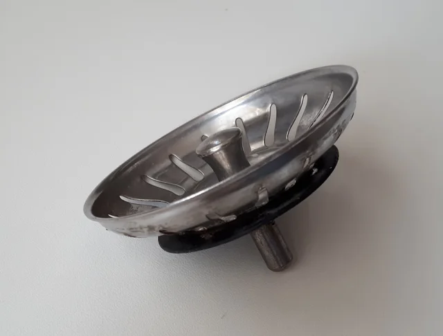Metal sink-strainer basket viewed from the side, with slotted walls, a central knob and a projecting stem underneath