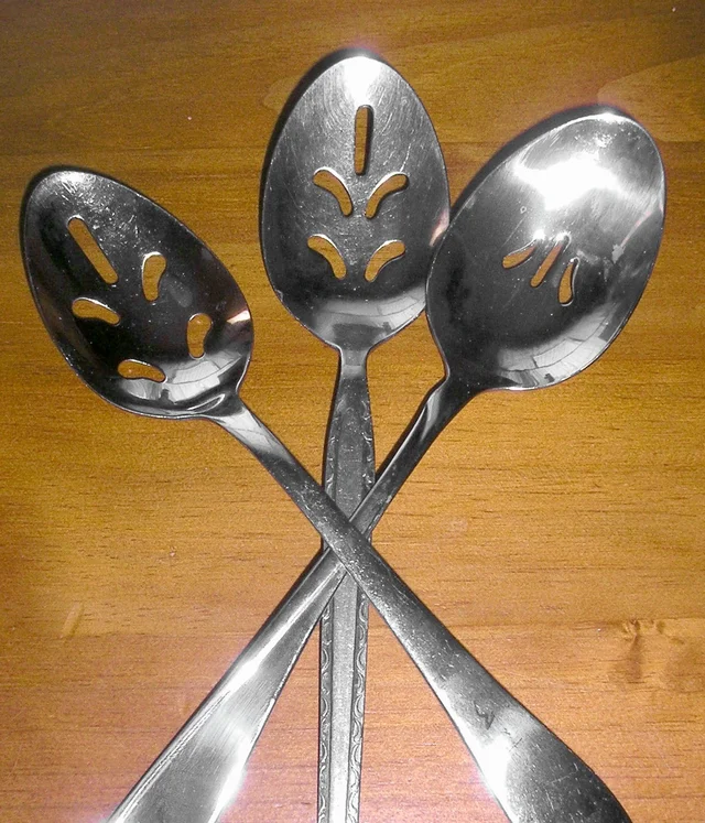 Three metal serving spoons with different slot patterns and crossed handles on a wooden surface