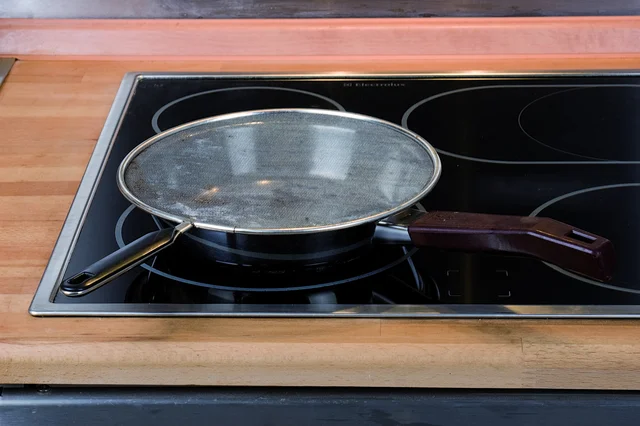 Round mesh splatter screen resting over a frying pan on a glass hob, with separate screen and pan handles